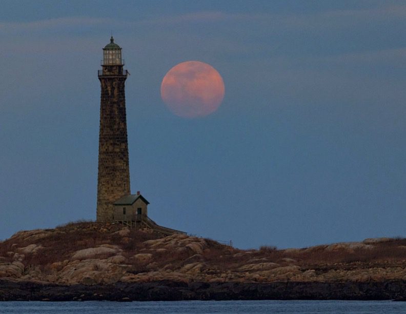 A lighthouse on the shore of an island with a full moon in the background.